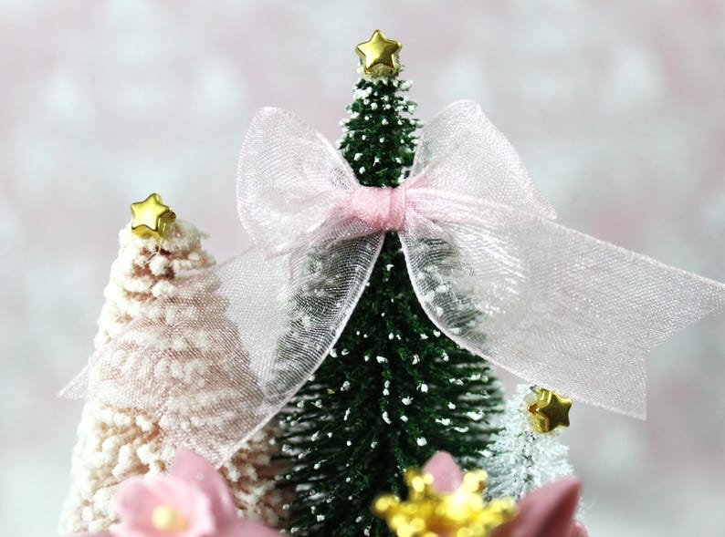 May include: Miniature Christmas trees, one green and two white, are decorated with gold star toppers and a sheer pink bow. The trees are adorned with faux snow and pink floral accents, creating a festive holiday scene.