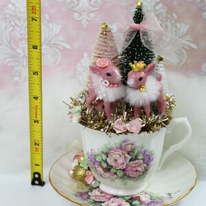 May include: A decorative teacup and saucer set with a floral design, holding a Christmas-themed arrangement. The arrangement includes two pink deer figurines, miniature trees, and gold tinsel. The height of the arrangement is approximately 8 inches.