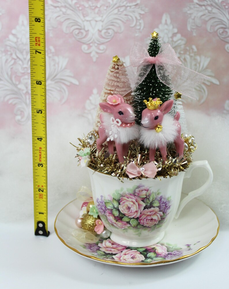 May include: A decorative teacup and saucer set with a floral design, holding a Christmas-themed arrangement. The arrangement includes two pink deer figurines, miniature trees, and gold tinsel. The height of the arrangement is approximately 8 inches.