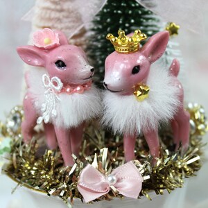 May include: Two pink deer figurines with white fur collars and decorative accents. One deer wears a flower, the other a gold crown. They stand on gold tinsel, with a pink bow and pearl detail.