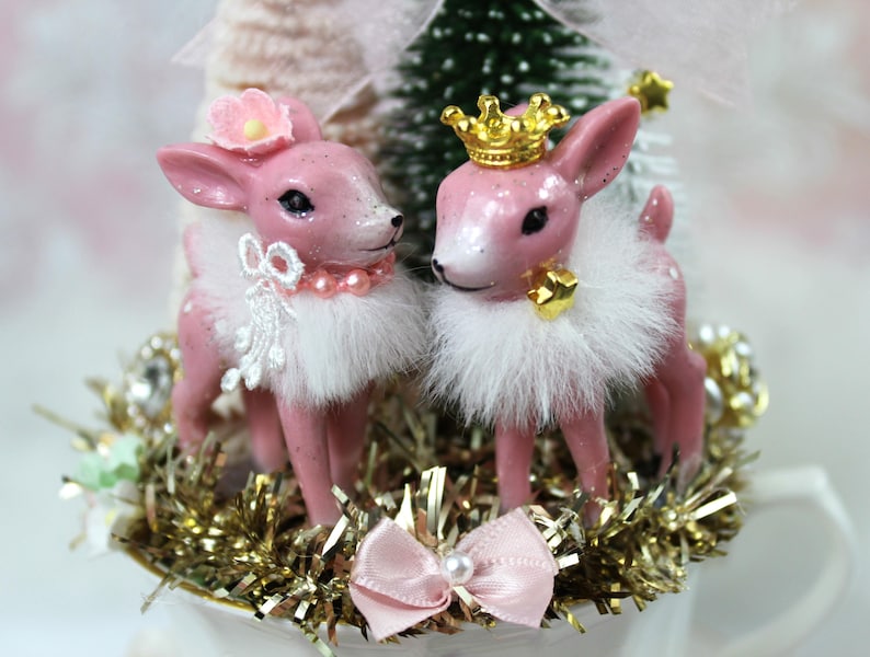 May include: Two pink deer figurines with white fur collars and decorative accents. One deer wears a flower, the other a gold crown. They stand on gold tinsel, with a pink bow and pearl detail.