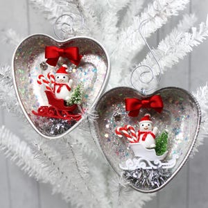 May include: Two heart-shaped Christmas decorations with silver glitter interiors. Each features a snowman in a red sleigh, candy canes, a small Christmas tree, and a red bow. The ornaments hang from silver hooks.