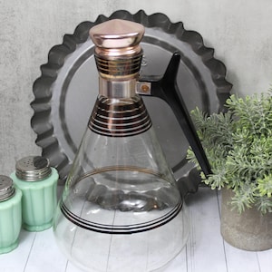 May include: A vintage glass soda siphon with a copper-colored top and black handle, alongside two mint green salt shakers with silver lids. A decorative metal plate and a small potted plant complete the arrangement.