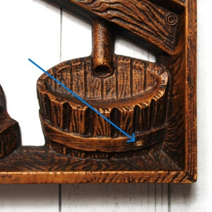 May include: A wooden wall decoration featuring a carved image of a well with a bucket. The well is made of dark brown wood with a light brown bucket. The bucket is hanging from a rope that is attached to a pulley.