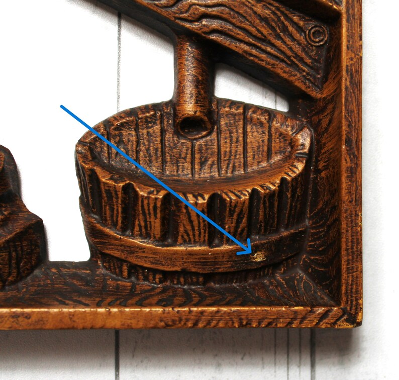 May include: A wooden wall decoration featuring a carved image of a well with a bucket. The well is made of dark brown wood with a light brown bucket. The bucket is hanging from a rope that is attached to a pulley.