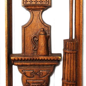 May include: A wooden wall decoration featuring a carved design of a stove, a pitcher, and a broom. The stove has a decorative design with a face and a row of teeth. The pitcher has a lid and a handle. The broom has a long handle and a bundle of straw at the bottom.
