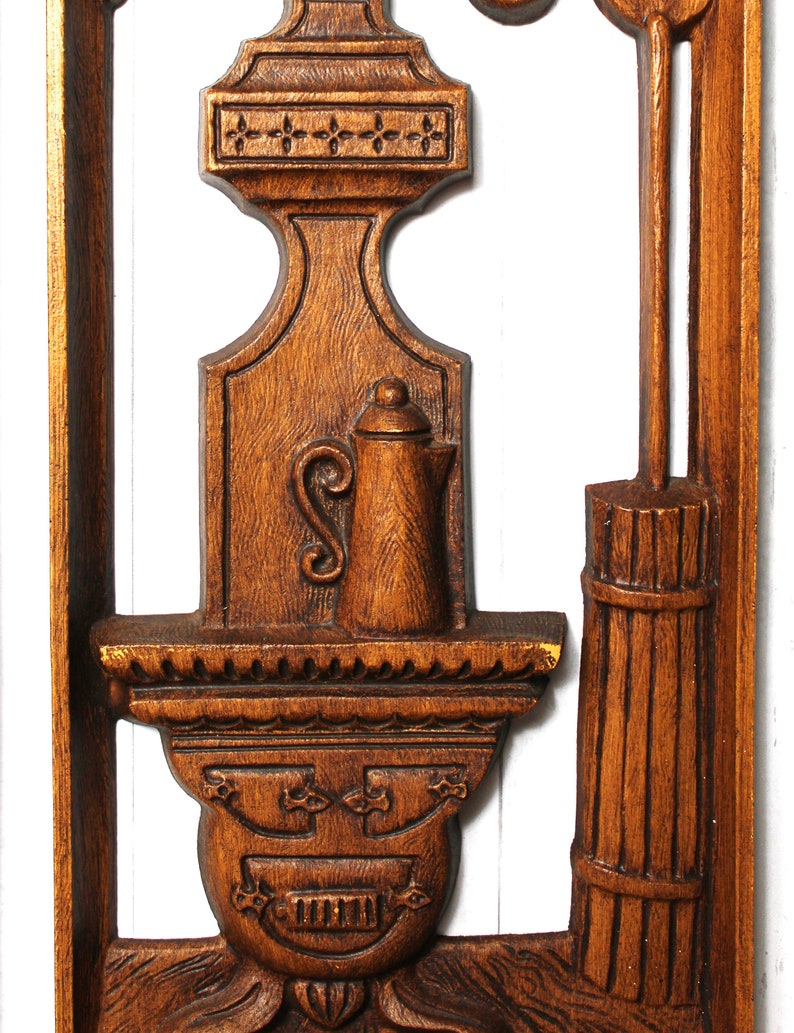 May include: A wooden wall decoration featuring a carved design of a stove, a pitcher, and a broom. The stove has a decorative design with a face and a row of teeth. The pitcher has a lid and a handle. The broom has a long handle and a bundle of straw at the bottom.