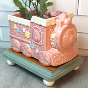 May include: Pink ceramic train planter with a teddy bear face and floral details. The planter is on a wooden base with white feet.