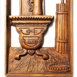 May include: A wooden wall decoration featuring a carved image of a stove with a shelf above it, a pitcher on the shelf, and a basket on the floor. The stove has a decorative design with a raised pattern and a small door. The wood is a rich brown colour.