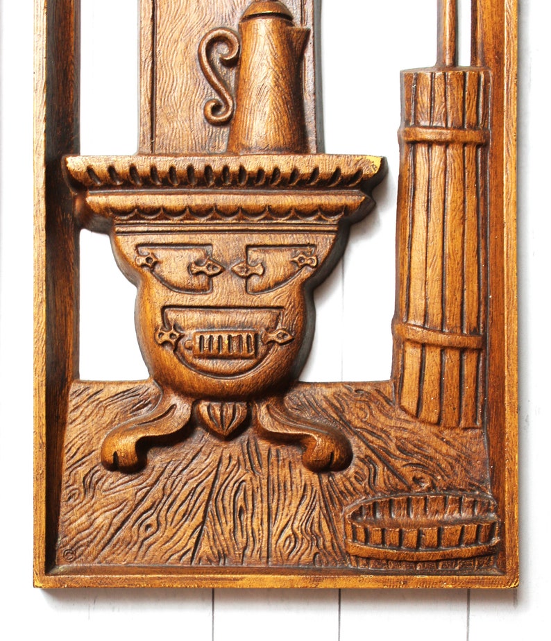 May include: A wooden wall decoration featuring a carved image of a stove with a shelf above it, a pitcher on the shelf, and a basket on the floor. The stove has a decorative design with a raised pattern and a small door. The wood is a rich brown colour.