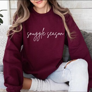 May include: Burgundy crewneck sweatshirt with the phrase "snuggle season" in white script. The long-sleeved top is made of a soft material. The sweatshirt is a comfortable and casual style.