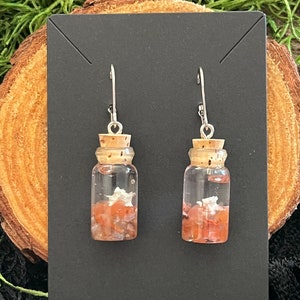 May include: A pair of silver earrings with small glass bottles filled with pink and white crystals. The bottles have cork tops and are suspended from silver hooks.