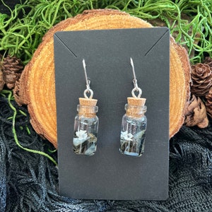 May include: A pair of silver dangle earrings with small glass bottles filled with black and white stones and a cork stopper.