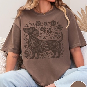 May include: Espresso-colored t-shirt featuring a dachshund dog design surrounded by floral elements. The dog and floral design are in a darker brown shade. The shirt is a relaxed fit and has a crew neck. The word "Espresso" is in a circle at the bottom right.