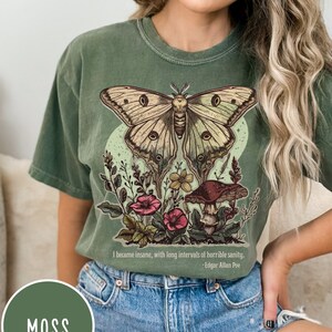 May include: A green t-shirt featuring a large, detailed illustration of a moth with large eyes, surrounded by flowers and mushrooms. The text "I became insane, with long intervals of horrible sanity. - Edgar Allan Poe" is printed below the illustration.