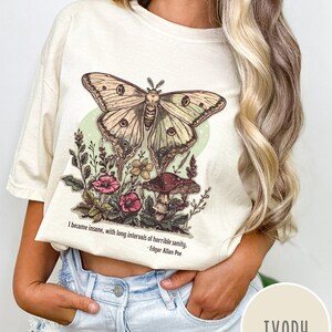 May include: A cream coloured t-shirt featuring a vintage illustration of a butterfly, mushrooms and flowers. The text "I became insane, with long intervals of horrible sanity. -Edgar Allan Poe" is printed on the shirt.