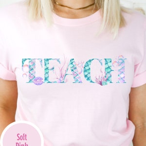 Puede incluir: Una camiseta rosa suave con la palabra "TEACH" escrita en letras blancas grandes. Las letras están decoradas con un patrón de escamas de sirena en tonos azules y verdes, con acentos rosados y burbujas.
