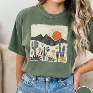 May include: Moss green t-shirt featuring a desert landscape design. The graphic includes a sun, mountains, and various cacti and desert plants. The shirt is paired with denim shorts.