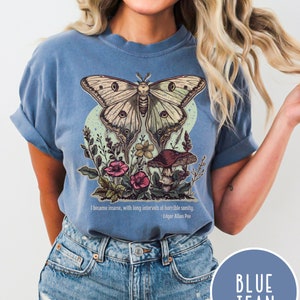May include: A blue t-shirt featuring a green and white butterfly with brown accents, surrounded by flowers and a mushroom. The text "I became insane, with long intervals of horrible sanity. - Edgar Allan Poe" is printed below the graphic.