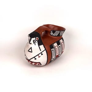 Può includere: Un vaso in ceramica dipinto a mano con un design unico. Il vaso presenta un manico e un bordo marrone, con un corpo bianco adornato da motivi geometrici neri, rossi e marroni. Il design include un volto stilizzato e forme geometriche.