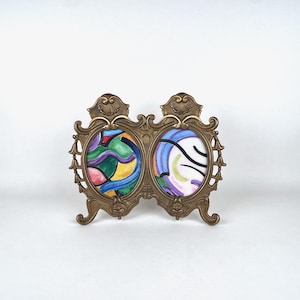 Puede incluir: Un marco de fotos doble ovalado hecho de metal en tono bronce antiguo con adornos de volutas y motivos de conchas. Las dos aberturas circulares contienen pinturas abstractas con remolinos vibrantes de colores verde, amarillo, azul, morado y rosa.