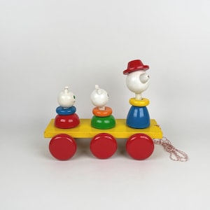 May include: A colorful wooden pull-along toy train with three figures and three red wheels. The figures are white with painted features, and the train has a yellow base. The toy is designed for young children.