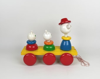 May include: A colorful wooden pull-along toy train with three figures and three red wheels. The figures are white with painted features, and the train has a yellow base. The toy is designed for young children.