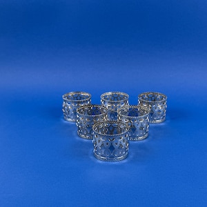 May include: Six silver napkin rings with an openwork design of connected diamond shapes. The napkin rings are arranged on a blue background. The rings are cylindrical and have a beaded edge.