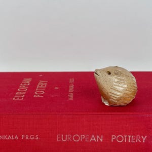 Puede incluir: Una pequeña figura de erizo de cerámica de color tostado con motas y líneas talladas que representan las púas, colocada sobre un libro rojo brillante titulado European Pottery de Maria Penkala F.R.G.S. La figura tiene un cuerpo redondeado.