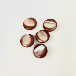 Puede incluir: Cinco botones redondos, marrón oscuro, con un diseño de media luna nacarada blanquecina. Los botones están dispuestos sobre una superficie blanca, mostrando su acabado brillante y sutiles variaciones de color. Ideales para costura y manualidades.
