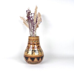 Könnte beinhalten: Eine beigefarbene Keramikvase mit rundem Boden und verjüngtem Hals, verziert mit handgemalten schwarzen geometrischen Mustern, horizontalen Linien und stilisierten Figuren. Die Vase enthält einen Strauß aus getrockneten Weizenähren und violetten Blumen.