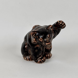 Könnte beinhalten: Eine Keramik-Bärenfigur, dunkelbraun mit hellbraunen Details, in sitzender Pose mit erhobener Pfote. Die Figur hat eine glänzende Oberfläche. Ein dekorativer Artikel.