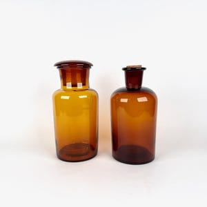 May include: Two amber glass apothecary bottles with cylindrical bodies and wide necks. The left bottle features a matching amber glass stopper, while the right bottle is topped with a small, light brown cork stopper. Both vessels are translucent.
