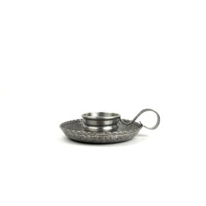 May include: A small, antique-style pewter candle holder. The candle holder has a round base with a textured pattern, a central cup for the candle, and a curved handle. The metal has a slightly tarnished appearance.