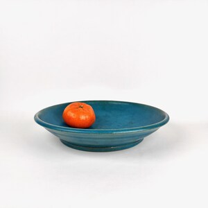 Puede incluir: Un cuenco de cerámica de color turquesa con un diseño poco profundo. Una mandarina naranja se encuentra en el centro del cuenco. El cuenco tiene una superficie texturizada y una base elevada. El fondo es blanco.