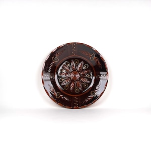 May include: A round, dark brown ceramic plate with a detailed floral design in the center. The plate features embossed patterns and a glossy finish. The rim has a decorative border with leaf-like shapes and small dots.