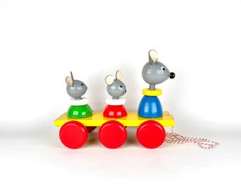 May include: A colorful wooden pull toy featuring three gray mice riding on a yellow cart with red wheels. The mice wear different colored outfits: green, red, and blue. A string is attached for pulling.