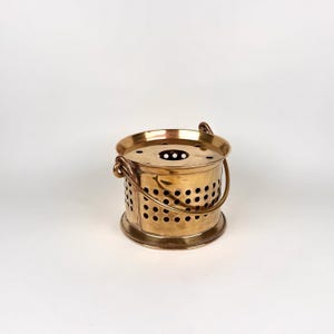 May include: A small, round, brass-colored bucket with a handle. The bucket has a series of evenly spaced holes around the sides and a circular top with a few small holes. The handle is attached to the sides with curved brackets.
