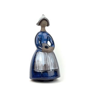 Può includere: Statuina in ceramica di una donna in abito tradizionale olandese. La statuina è principalmente blu con un grembiule e un cappello bianchi. La donna tiene in mano un piccolo oggetto. La statuina è in piedi.