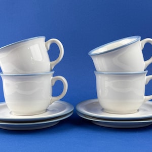 May include: Four white ceramic teacups and saucers with a light blue trim. The cups are stacked in pairs, each set on a matching saucer. The background is a solid blue.