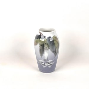 May include: A small, decorative vase with a floral design. The vase features a light blue gradient, transitioning to white at the top. The painted design includes green and grey leaves and a white flower. The vase is approximately 15 cm tall.