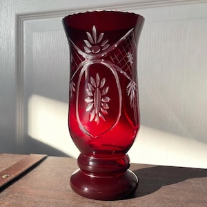 Può includere: Un vaso di vetro rosso rubino con intricati disegni floreali e geometrici incisi. Il vaso ha una parte superiore svasata, un corpo cilindrico e una base arrotondata. I motivi incisi includono fiori stilizzati, stelle e tratteggio incrociato.