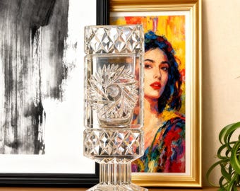 Puede incluir: Un jarrón de cristal tallado transparente con un diseño de estrella se encuentra sobre un mueble de madera. Una pintura abstracta en blanco y negro y un retrato enmarcado de una mujer están detrás del jarrón. A la derecha hay una planta en maceta con hojas verdes largas.
