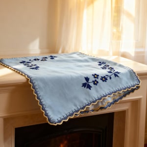 Może przedstawiać: Jasnoniebieski dekoracyjny bieżnik na stół z haftowanymi ciemnoniebieskimi wzorami kwiatowymi. Bieżnik ma falisty brzeg ze złotym wykończeniem. Prezentowany jest na białym kominku.