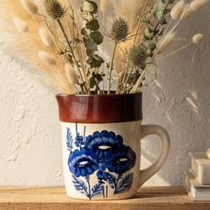 Può includere: Un boccale in ceramica con bordo marrone e motivo floreale blu contiene un bouquet di fiori secchi, tra cui erba della pampa, cardi ed eucalipto. Il boccale è su una superficie di legno, con ombre sul muro.