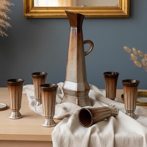 May include: A vintage ceramic jug and glasses set. The tall jug has a brown top and handle, with a light brown and beige body. Six matching glasses with brown rims and bases are arranged around the jug on a linen cloth.