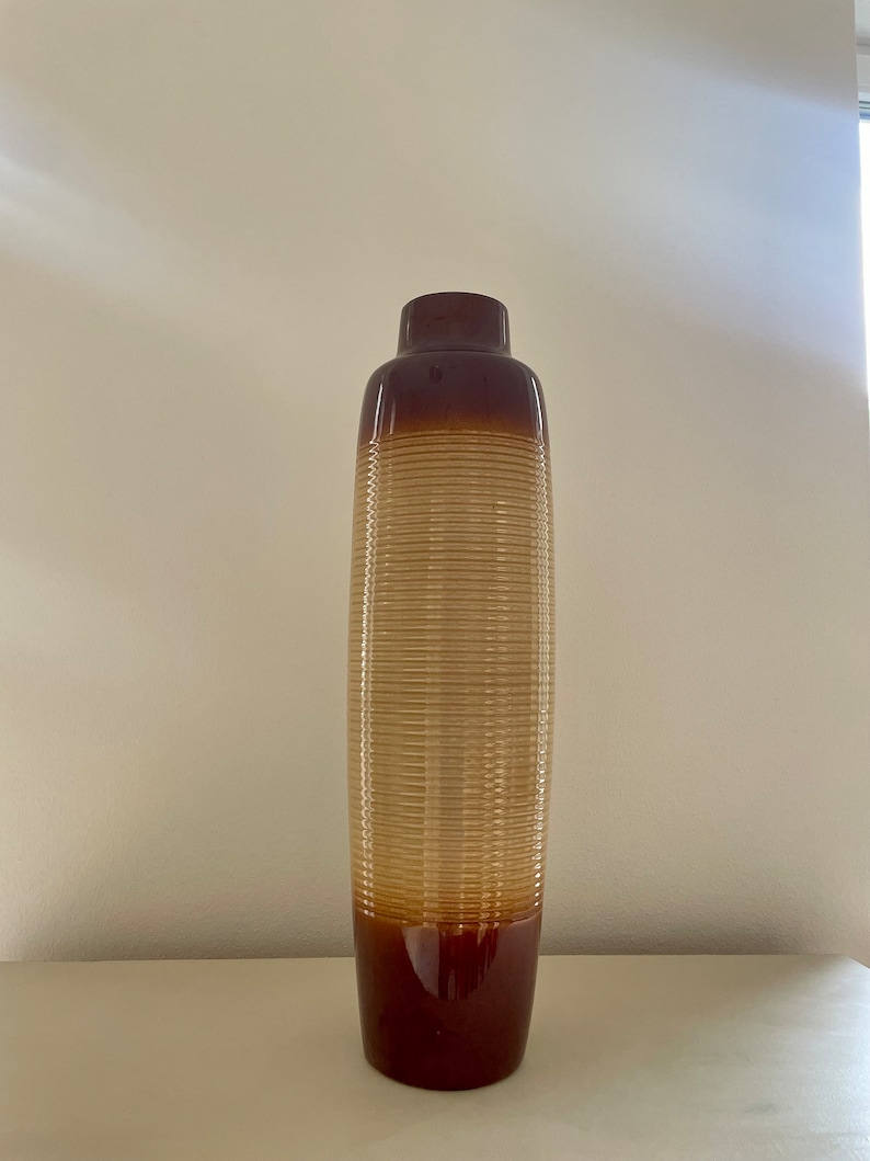 May include: A tall, decorative vase with a cylindrical shape. The vase features a gradient of colors, transitioning from a dark brown base to a lighter tan center with horizontal ribbed detailing, and a dark brown top.