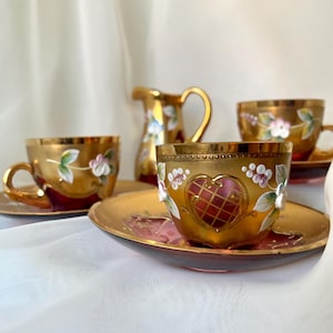May include: A gold-colored tea set with floral and heart designs. The set includes four teacups with saucers and a small creamer. The cups and creamer have gold rims and handles. The floral designs are in white and pink.