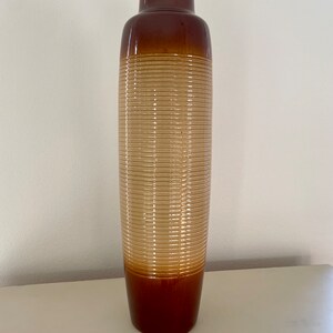 May include: A tall, brown ceramic vase with a cylindrical shape. The vase features a ribbed texture in a light tan color in the middle, with a darker brown top and base. The vase is approximately 24 inches tall.