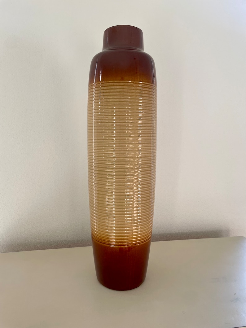May include: A tall, brown ceramic vase with a cylindrical shape. The vase features a ribbed texture in a light tan color in the middle, with a darker brown top and base. The vase is approximately 24 inches tall.